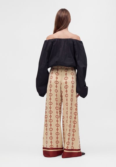 Femme aux longs cheveux porte un top noir épaules dénudées avec des manches surdimensionnées et un pantalon beige à jambes larges avec un motif chaîne rouge, debout de dos.