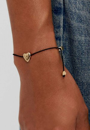 Bracelet en cordon noir avec un charme en forme de cœur doré et un fermoir coulissant. Le cœur est stylisé avec une texture lisse et accompagné d'un petit cœur en accent.