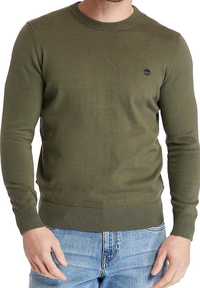 Homme portant un pull col rond vert olive avec un petit logo noir sur la poitrine et un jean bleu clair, debout, mains détendues.