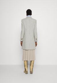 Lång rutig blazer med ett ljusgrått och svart rutig mönster, kombinerad med en beige veckad kjol och beige stövlar med tigerrandigt mönster.