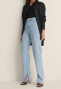 Vaalean siniset korkeavyötäröiset farkut, joissa on suora malli, etupuolella halkiot ja klassinen viiden taskun design, yhdistettynä mustaan nappipaitaan.