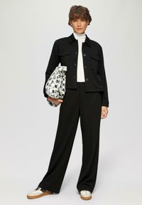 Giacca nera cropped con chiusura a bottoni e due tasche frontali, abbinata a pantaloni neri a vita alta e un dolcevita bianco. Tiene in mano una borsa con motivo.