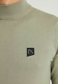 Pull en maille côtelée vert clair avec un col haut. Présente un patch carré noir avec un logo blanc sur la poitrine. Texture douce.