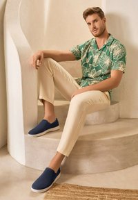 Granatowe buty wsuwane, teksturowany materiał, biała podeszwa; jasnobeżowe lniane spodnie, koszula w zielony tropikalny wzór, siedzący na zakrzywionych schodach.