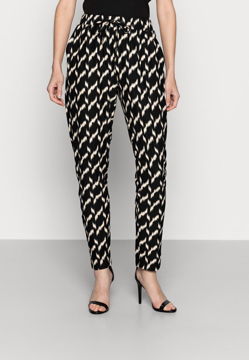 Femme portant un pantalon à motifs zigzag noir et beige avec une taille élastique et des sandales noires à talons hauts, debout devant un fond uni.