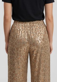 Persona con pantalones brillantes de estampado de leopardo dorado con cinturilla elástica y una camiseta negra lisa de manga corta, vista desde atrás.