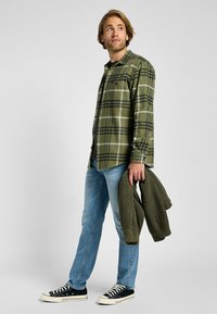 Grön rutig flanellskjorta med svarta och vita detaljer, bärs över en svart tröja; kombineras med ljusblå jeans och svarta sneakers.