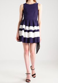 Laivansininen hihaton mekko, jossa on laskostettu hame ja valkoisia vaakaraitoja. Korostamassa musta käsilaukku ja mustat nauhakengät.