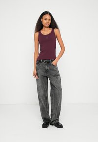 Vīnogu krāsas bezroku tops ar logotipu, kombinēts ar vaļīgām, izbalējušām melnām džinsu biksēm un melnām sandālēm, radot ikdienišķu apģērbu.