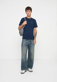 Dunkelblauses Baumwoll-T-Shirt mit Rundhalsausschnitt, kombiniert mit hellblauen weit geschnittenen Jeans. Graue Umhängetasche und weiße Sneaker mit schwarzen Akzenten.