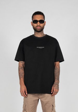 Black cotton t-shirt with loose fit, short sleeves, and a white printed logo in the center. Worn with beige cargo pants and sunglasses.
