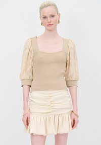 Top beige in maglia con maniche a sbuffo in pizzo, scollo quadrato e design aderente. Abbinato a una minigonna crema arricciata con orlo a volant.