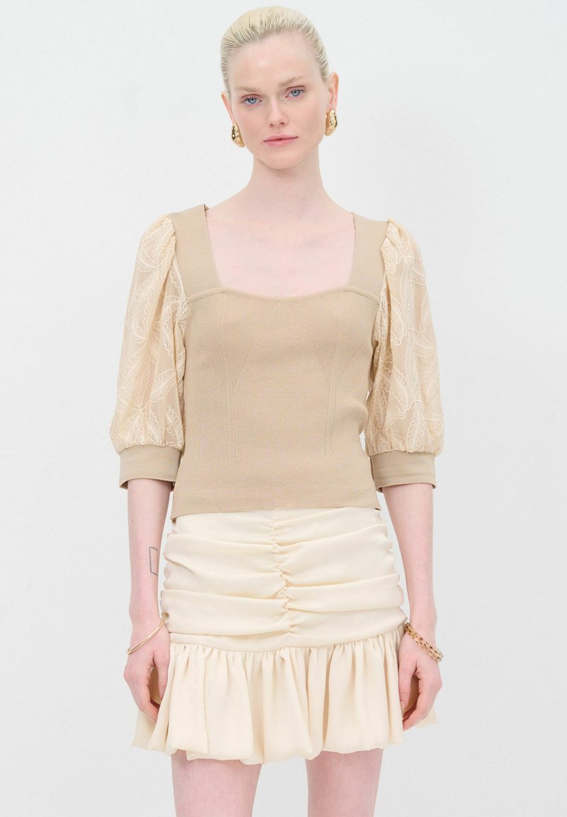 Beige gebreide top met kanten pofmouwen, vierkante halslijn en aansluitend ontwerp. Gecombineerd met een crèmekleurige mini rok met ruches en een gerimpelde zoom.