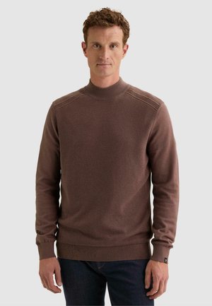 Hombre con cabello corto y rizado que lleva un suéter marrón de punto de manga larga y pantalones oscuros, de pie frente a un fondo claro y sencillo.