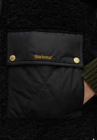 Gros plan d’une poche de veste matelassée noire Barbour avec des pressions dorées et une main partiellement dans la poche.
