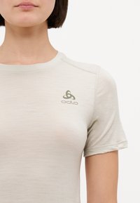 Gai smilšu krāsas, piegulošs Odlo t-krekls ar īsām piedurknēm, gluda tekstūra un neliels tumši zaļš logotips augšējā kreisajā stūrī. Mīksts, minimālisma dizains.