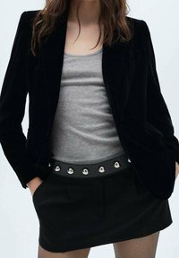 Blazer en velours noir porté sur un débardeur gris, associé à une jupe noire. La jupe se distingue par une ceinture en cuir ornée de clous métalliques.