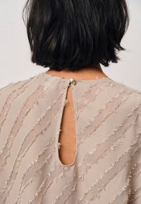 Beige Bluse aus leichtem Stoff, mit vertikalen Fransen, einem Schlüsselloch-Ausschnitt hinten und einem runden Knopfverschluss.