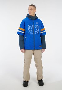 Blaue Mesh-Jacke mit orange und weißen Streifen, auf der Vorderseite mit einer großen Nummer "93" versehen, kombiniert mit beigen Hosen und schwarzen Stiefeln.