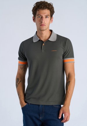 Giovane uomo con capelli ricci che indossa una polo grigio scuro con maniche rigate grigie e arancioni e jeans blu, in piedi con una mano in tasca.