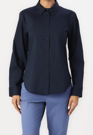 Mujer con una camisa azul marino ajustada de manga larga abotonada, metida dentro de unos pantalones azul claro, de pie contra un fondo liso.