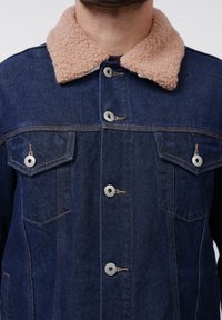 Chaqueta de denim con un cuello mullido de color marrón claro. Presenta botones plateados, dos bolsillos en el pecho y una textura de tela azul oscura.