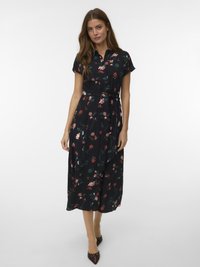 Γυναίκα που φοράει ένα μαύρο midi φόρεμα με λουλούδια, κοντά μανίκια, γιακά και ζώνη στη μέση, σε συνδυασμό με καφέ γόβες με μυτερή μύτη.