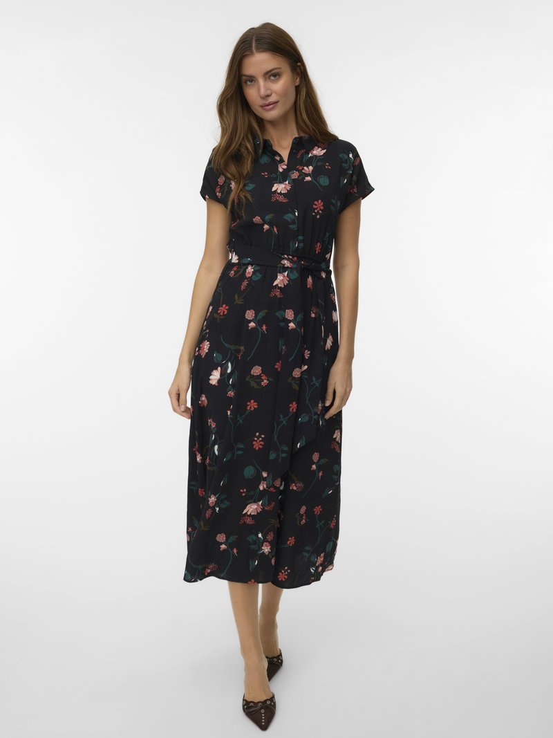 Γυναίκα που φοράει ένα μαύρο midi φόρεμα με λουλούδια, κοντά μανίκια, γιακά και ζώνη στη μέση, σε συνδυασμό με καφέ γόβες με μυτερή μύτη.