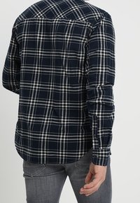 Camicia di flanella blu navy con motivo a quadretti bianchi e grigio chiaro, maniche lunghe e polsini con bottoni. Presenta un design a yoke e un orlo curvo.