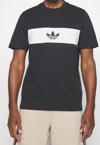 Černé bavlněné tričko s bílým vodorovným pruhm a vyšívaným logem uprostřed. Krátké rukávy, kulatý výstřih.