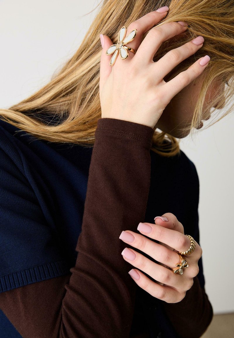 Donna con lunghi capelli biondi che indossa maniche marroni e blu scuro, mostrando anelli dorati a forma di fiore e foglia sulle dita e unghie curate.