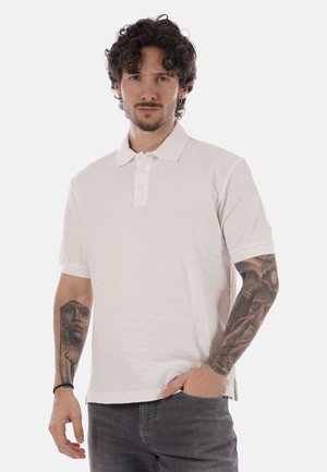 Mężczyzna z kręconymi włosami i kolczykami, ubrany w białą koszulkę polo i ciemne dżinsy, pokazujący szczegółowe tatuaże na obu przedramionach na białym tle.