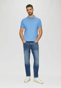 Hellblaues Polohemd mit Kragen, kurzen Ärmeln und kleinem Logo. Kombiniert mit blauen, verblassten Jeans und weißen Sneakers. Einfaches Design.