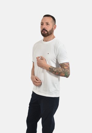 Uomo con capelli corti e barba che indossa una camicia bianca a maniche corte e pantaloni scuri, mostrando un tatuaggio floreale sulla manica del braccio destro, in piedi su sfondo bianco.