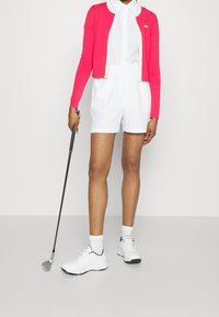 En ljusrosa kavaj över en vit skjorta med krage och vita shorts. Vita sportskor och en golfklubba hålls i ena handen.