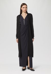 Czarny długi kardigan na ciemnoniebieskiej sukience z krojem kopertowym, z głębokim dekoltem w kształcie V i bocznym rozcięciem, zestawiony z czarnymi balerinami.