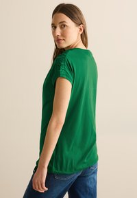 Grøn kortærmet T-shirt lavet af letvægtsstof, med rynkede detaljer på skuldrene og en afslappet pasform, parret med mørkeblå jeans.