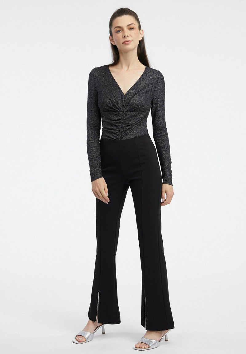 Schwarzes Longsleeve-Oberteil mit einem verdrehten Frontdetail, kombiniert mit engen schwarzen Hosen mit seitlichen Schlitzen und silbernen High Heels.