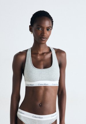 Siva pamučna sportska grudnjak s širokim bijelim elastičnim pojasom na kojem se nalazi logo "Calvin Klein". Model nosi odgovarajuće sive gaćice.