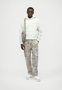 Helle graue Kapuzenjacke mit einer Fronttasche, kombiniert mit beigen, camo-musterierten Cargo-Hosen und weißen Sneakers. Akzentuiert mit einer grauen Umhängetasche.