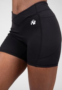 Zwarte sportieve shorts van rekbaar materiaal, met vlakke naden en een wit logo aan de linkerkant. Korte lengte en aansluitend ontwerp.