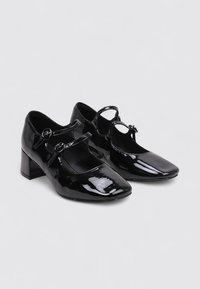Chaussures Mary Jane en cuir verni noir avec deux sangles à boucle et de petits talons blocs sur un fond gris clair uni.