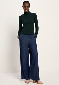 Frau steht mit den Händen in den Taschen, trägt einen dunkelgrünen Rollkragenpullover, weite blaue Jeans und rosa Turnschuhe vor einem einfarbigen Hintergrund.