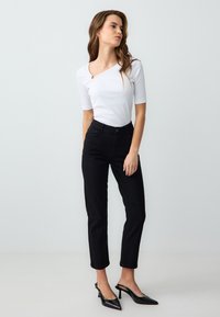 Top bianco a coste con scollatura curva, arricchito da un dettaglio anello dorato, abbinato a pantaloni neri a vita alta e tacchi a punta neri.