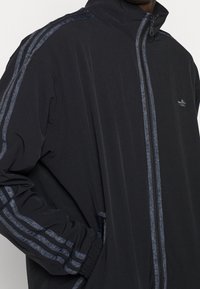 Man som bär en svart zip-up jacka med mörkgrå ränder längs ärmarna och dragkedjan, logotyp på bröstet, hand i sidofickan.