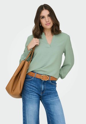 Frau trägt eine hellgrüne langärmelige Bluse, blaue Jeans mit einem hellbraunen Gürtel und trägt eine große hellbraune Ledertasche über der Schulter.