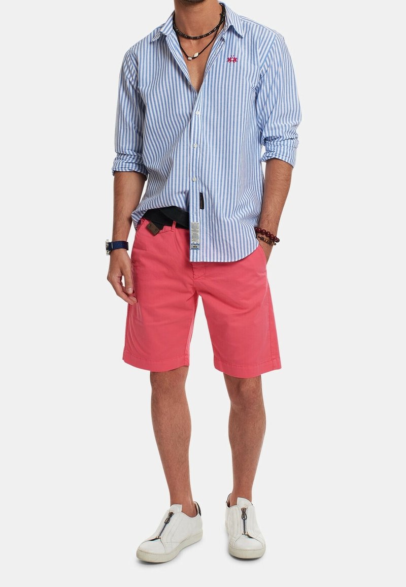 Striped blue button-up shirt, pink shorts, white sneakers. Details include a logo on the shirt and mixed accessories like necklaces and bracelets.