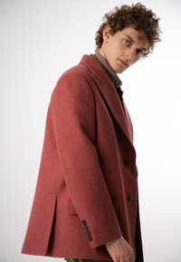 Czerwony wełniany płaszcz z klapami, podwójnie zapięty z przodu, z bocznymi kieszeniami i czarnymi guzikami. Gładka tekstura i zdefiniowana sylwetka.