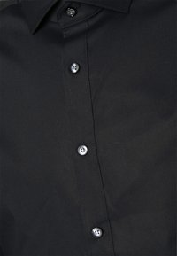Chemise noire à boutons avec un col classique, en tissu lisse et des boutons ronds noirs à l'aspect légèrement brillant.