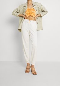 Modelka pózuje v nabraném oranžovém crop topu, béžové oversized bundě a bílých joggingových kalhotách, doplněná hnědými sandály na podpatku na neutrálním pozadí.